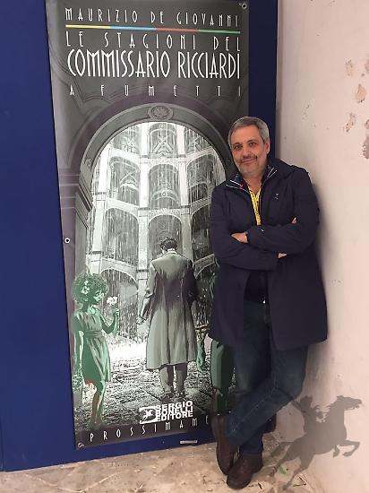 Maurizio De Giovanni posa accanto al cartellone che annuncia "Le stagioni del Commissario Ricciardi" a fumetti.